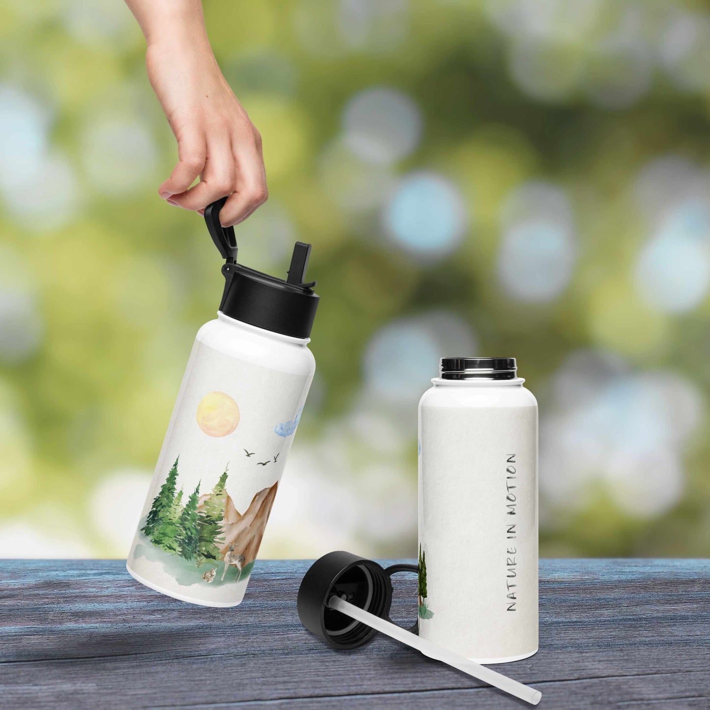 White water bottle with nature-themed illustration of a forest scene with trees, mountain, rocks, and animals on a wooden surface with a blurred green background.