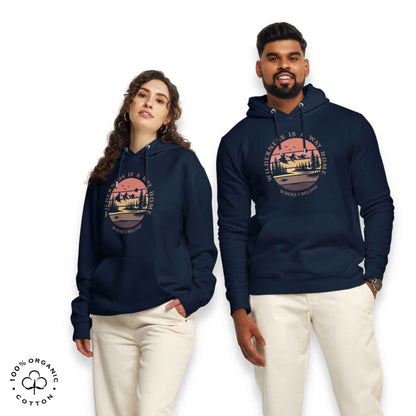 Woman and men wearing navy organic cotton hoodie with nature-themed graphic, mountain, forest, sun and river. With bold Wilderness is a way home, where I belong. 